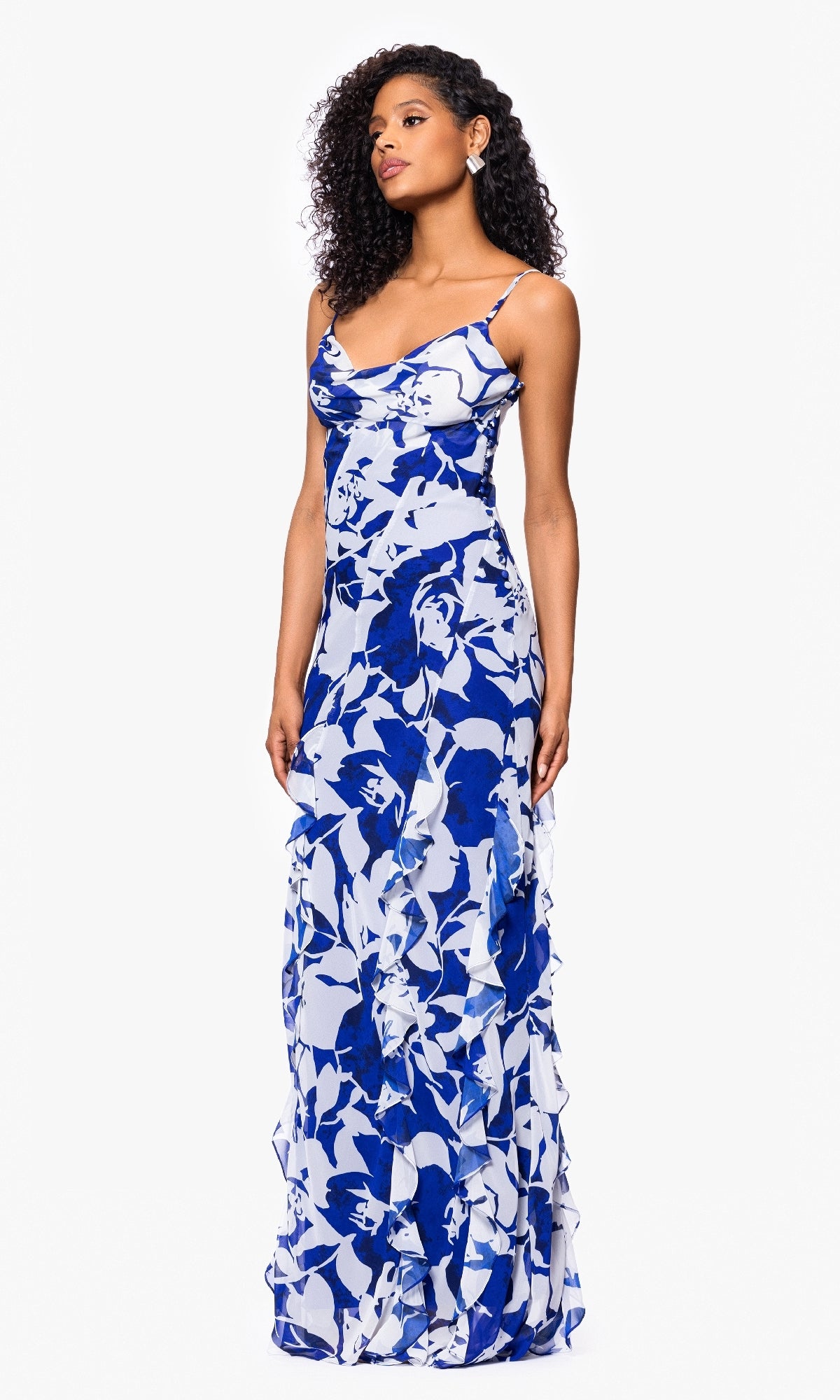 A woman wears the Betsy and Adam A26915 long formal dress, featuring a sleeveless blue and white floral print with subtle ruffle details, standing against a plain white background.